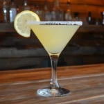 A vibrant lemon drop cocktail in a sugar-rimmed martini glass with a lemon slice garnish on the side, placed on a wooden bar counter.